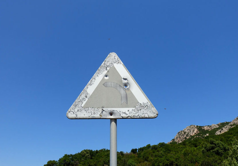 An old road sign on a mountain road indicating a dangerous curve, perhaps riddled with rifle shots.